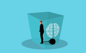 A man inside a cube. He is held back by a ball and chain. There is an image of a brain in the box with the man.